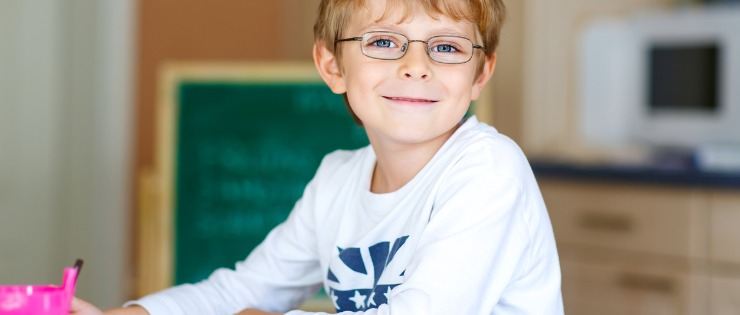 A school aged boy wearing glasses and doing homework at the kitchen table.