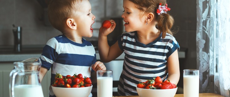 Two children feeding each other strawberries and fruit at the breakfast table.