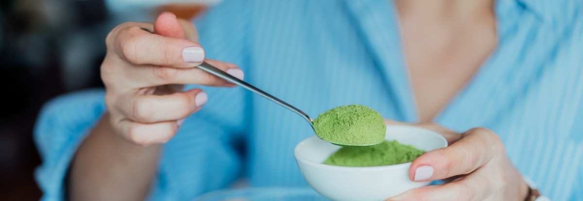 Close up of a woman adding a spoonful of a superfood green powder into a smoothie blender.