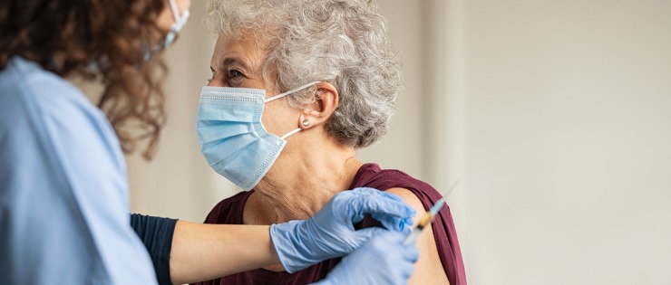A seated mature lady wearing a mask with her sleeve rolled up about to receive a vaccination administered by a nurse in scrubs.