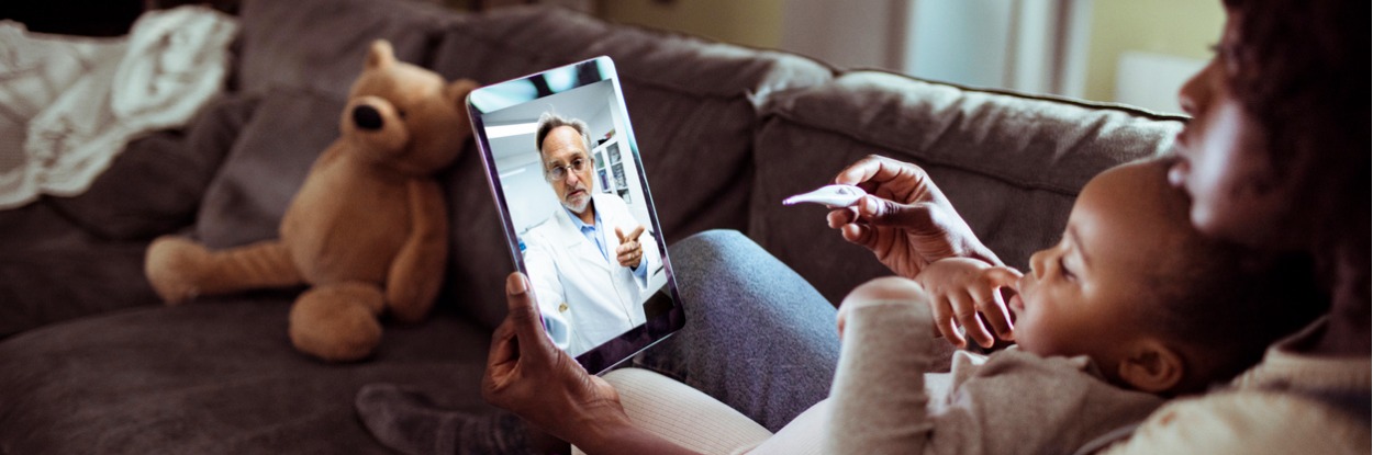 Woman sitting on the couch with her baby, showing the doctor a thermometer on a telehealth video conferencing appointment